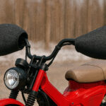 冬の屋外で赤い小型オフロードバイクのハンドル周りをクローズアップした写真風イメージ。黒いハンドルカバーが左右に装着され、背景には雪の残る林がぼんやりと広がっている。バイクの車体にはブランド名やロゴが一切なく、無地のデザインとして描かれている。