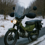 雪の残る冬の林道に停められたグリーンのバイク。ウインドシールドとハンドルカバーを装着した冬仕様カスタムのバイクが、寒冷な景色の中でライトを点灯させている写真。