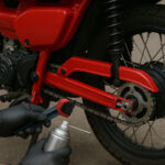 赤いデュアルスポーツバイクの後輪まわりで、チェーンをブラシとスプレーでメンテナンスしている様子が写っている。