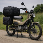 ツーリングバッグを装着した架空デザインのデュアルパーパスバイクが、草地のそばで撮影されている写真