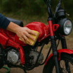 赤いオフロード風バイクを屋外で手洗いしている様子。スポンジでタンク部分を優しくこすり、水滴が車体に残っている。ヘッドライトやエンジン周りも濡れており、洗車作業のリアルな質感が伝わる写真。