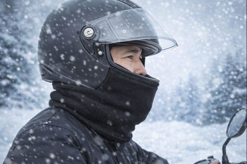 雪が降る冬の屋外で、黒いヘルメットとネックウォーマーを着用した男性ライダーがバイクに乗っている様子